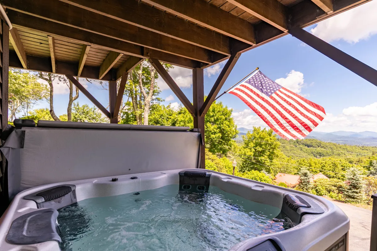 This image features a hot tub with a view of the mountains, the American flag in the background, and an inviting deck setting. The hot tub is front and center, emphasizing relaxation while soaking in natural beauty. The overall scene is tranquil and social, ideal for unwinding or gathering with friends and family.