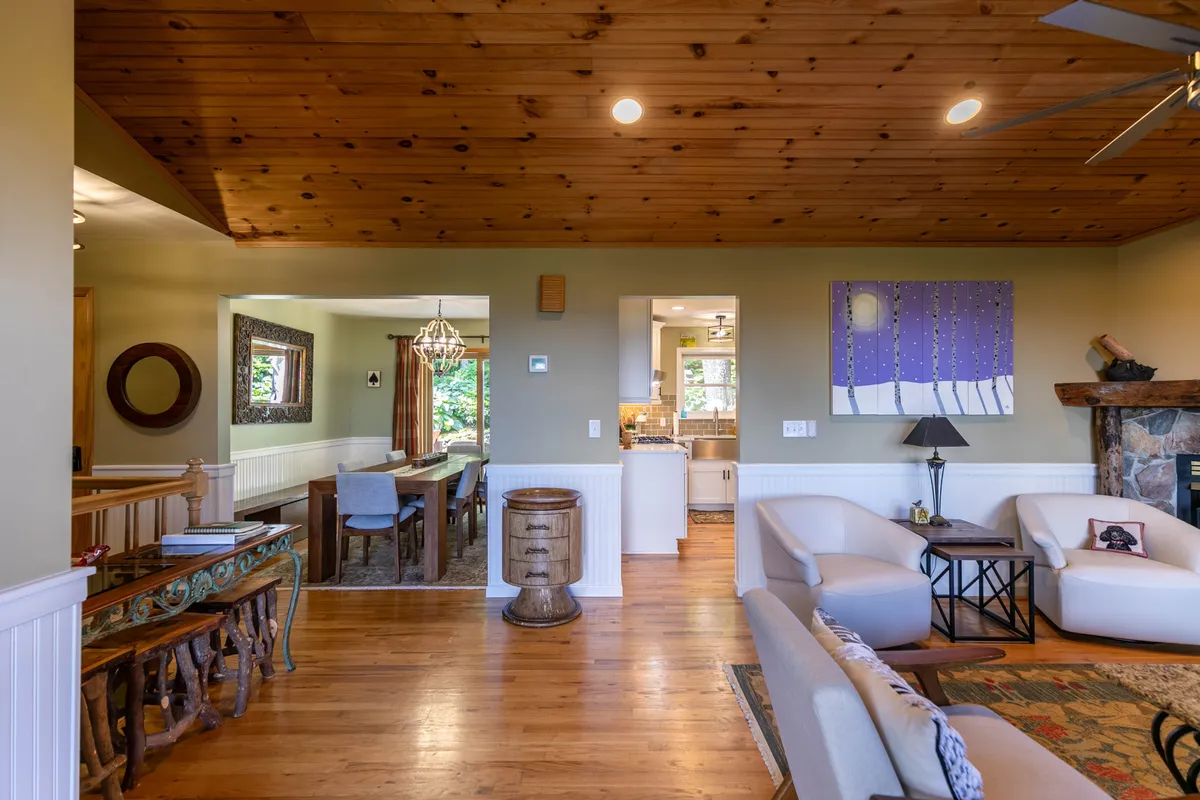 An open-concept space with a vaulted wooden ceiling, cozy dining area for ten, a glowing fireplace, and a glimpse of an adjoining kitchen. Artwork and comfortable seating create a welcoming environment ideal for large gatherings or family meals.
