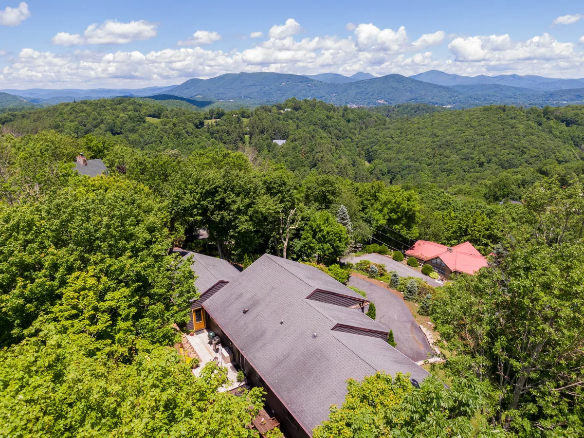 This image shows the lodge nestled against a stunning mountain backdrop, surrounded by tall trees. The house is visible with a deck area that provides direct access to the outdoors, highlighting the property's proximity to natural adventures.