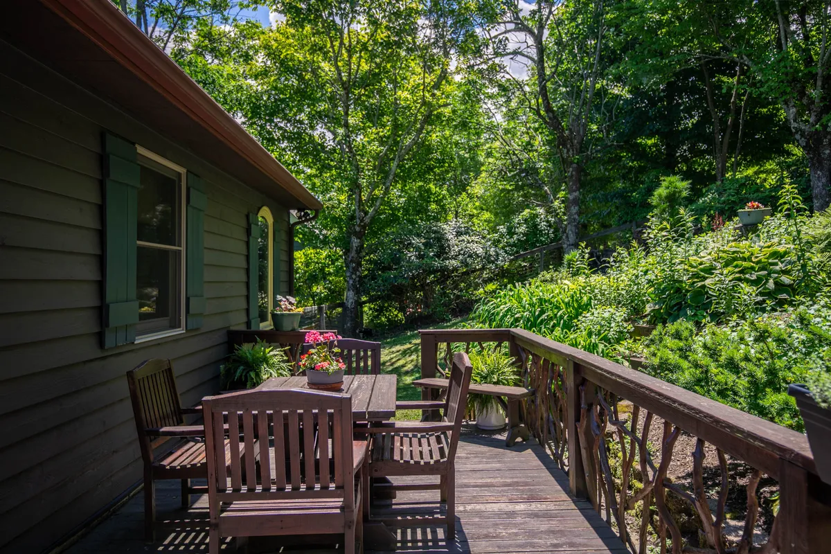 A charming outdoor dining area on a wooden deck, surrounded by lush greenery and flower pots. A table and chairs are set up to enjoy meals al fresco in a tranquil, natural setting—perfect for gatherings and sunset dinners.