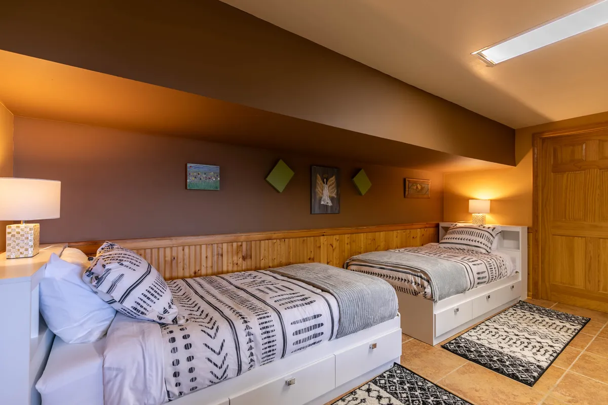 A cozy room with two twin beds, decorative lamp, wooden paneling, and tasteful artwork adorning the walls. Textile patterns on the bedding add warmth and a playful touch, making it ideal for children or teens.
