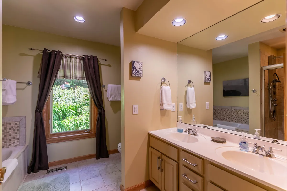 A spa-inspired bathroom with a double vanity, soaking bathtub, large window filling the space with morning light, and a glass shower enclosure. Sleek finishes and serene styling evoke a luxurious, restful ambiance.