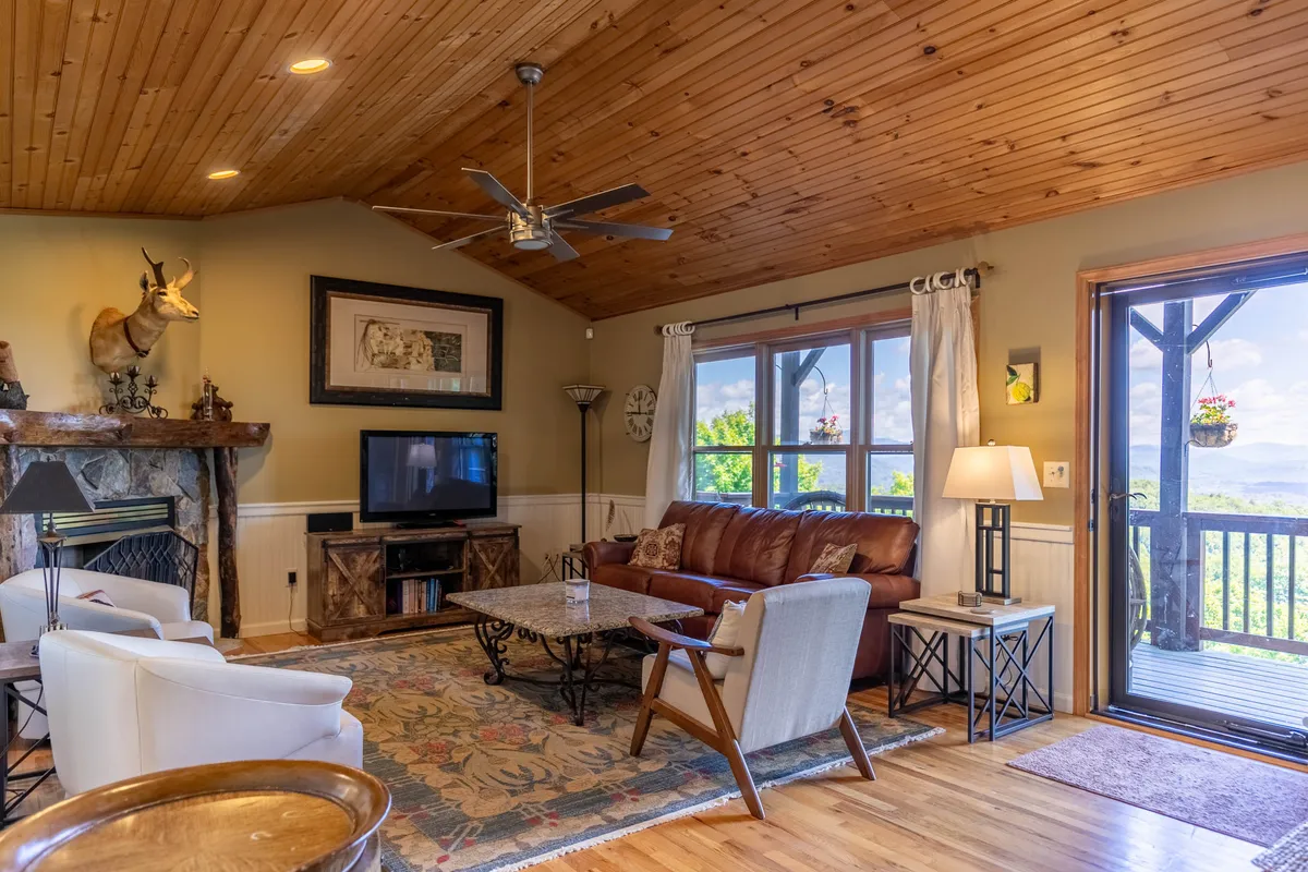 A spacious family room featuring a stone gas fireplace as the focal point, with a comfortable leather sofa arranged to enjoy the warmth and ambiance. Large windows frame stunning mountain vistas, filling the space with natural light. Decorative wall art and wood accents enhance the mountain lodge feel.
