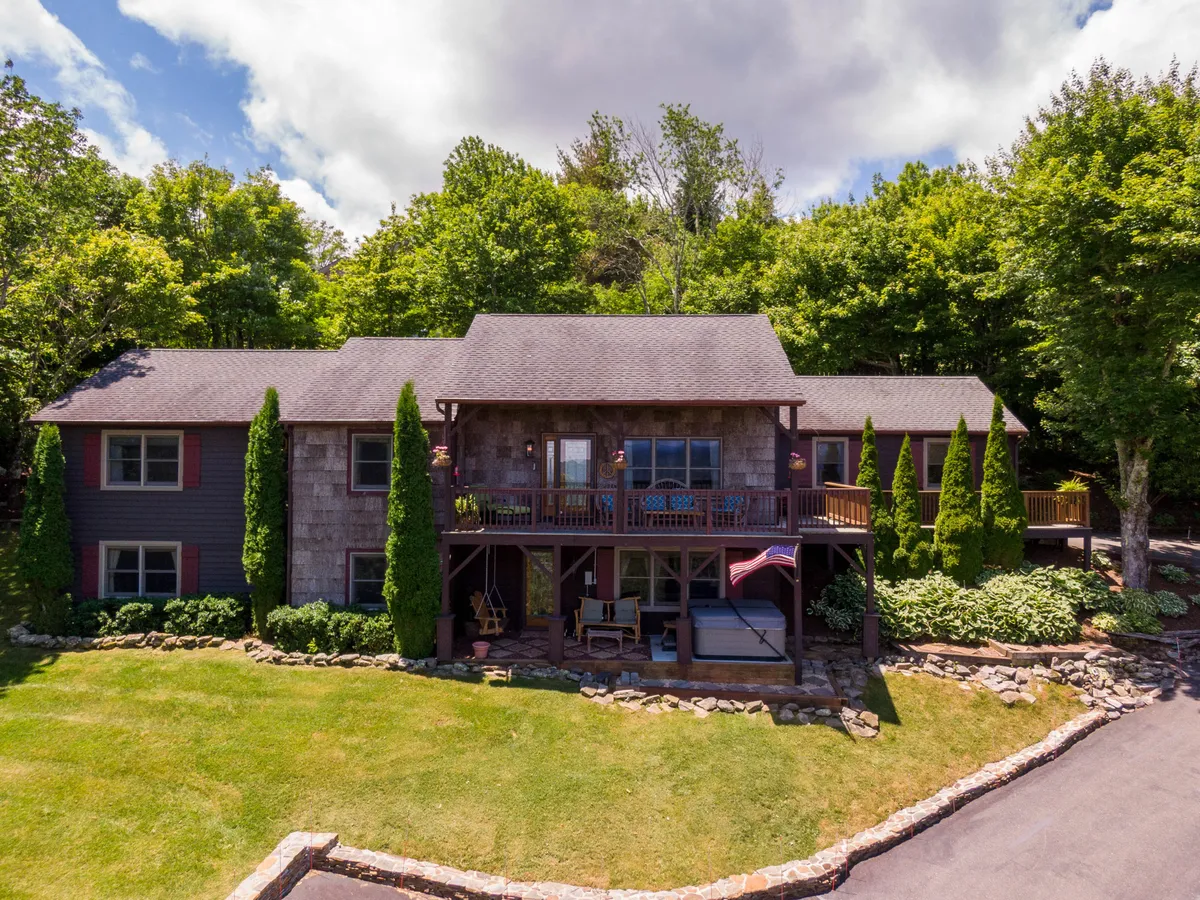 The image showcases the inviting front porch of a charming mountain home nestled among lush greenery. Large windows reflect the natural light, and a stone pathway leads up to the welcoming entrance. An American flag flutters, adding a touch of warmth and community. The scene exudes comfort and the promise of relaxing evenings spent enjoying breathtaking sunsets or family gatherings on the porch.