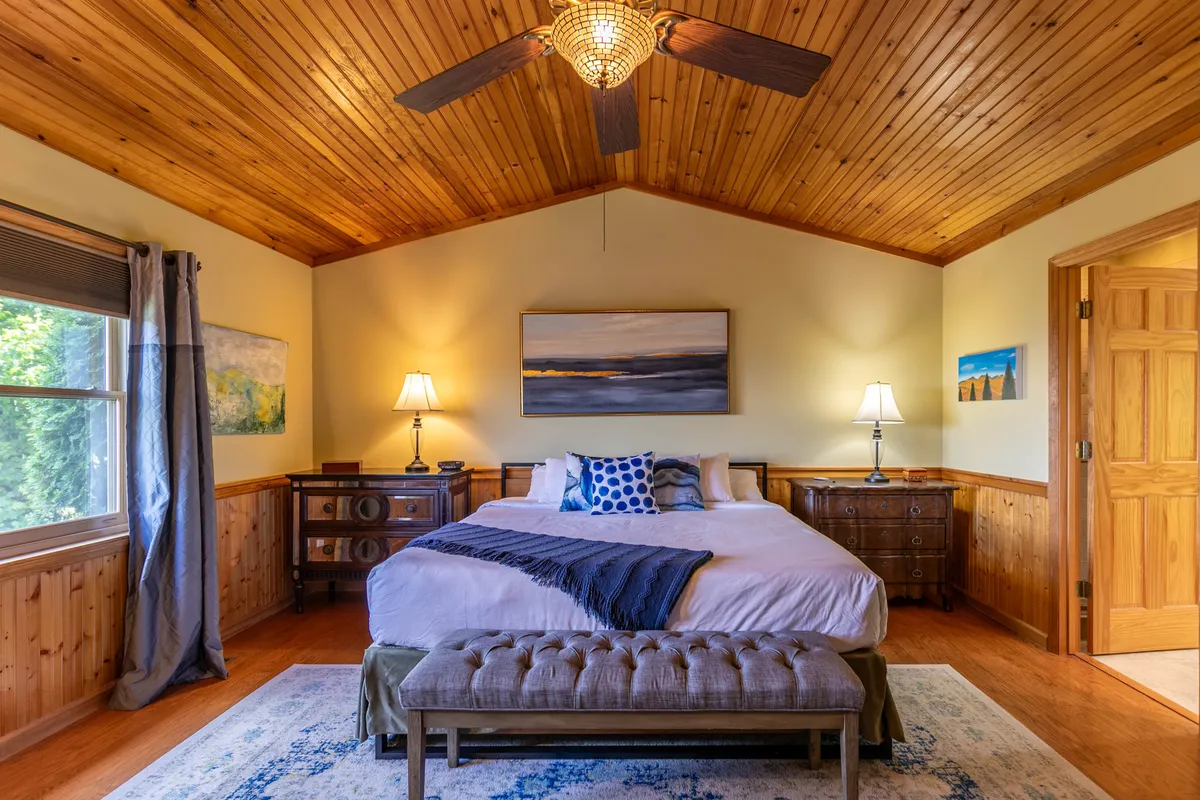 A tranquil bedroom featuring a plush bed with crisp linens, nightstands with lamps on either side, and artwork on the wall. A window with soft curtains lets in gentle natural light, creating a serene and inviting atmosphere.